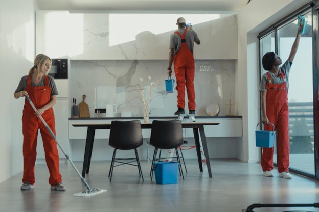 Professional cleaners in red overalls sanitizing a modern office space, mopping the floor, cleaning windows, and dusting surfaces, emphasizing the importance of cleanliness in workplaces.