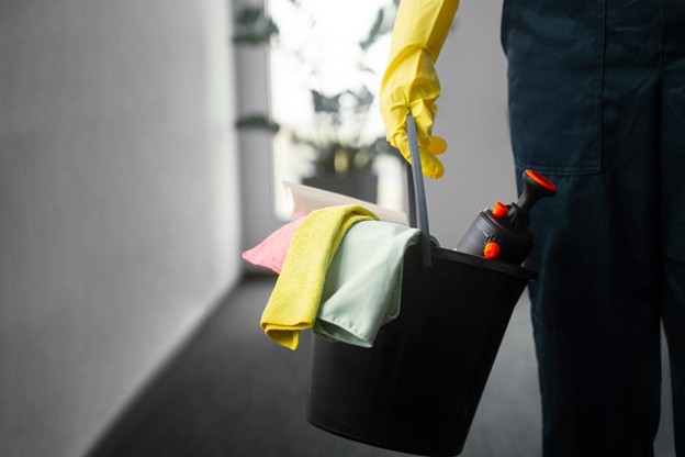 Cleaning professional holding a bucket with cleaning supplies, including colorful cloths and a spray bottle, in a well-lit office hallway, emphasizing the importance of thorough post-construction cleaning for a safe work environment.