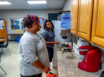 Two Clean Impact LLC team members engaging in conversation by a coffee station, emphasizing camaraderie and teamwork in a professional setting.